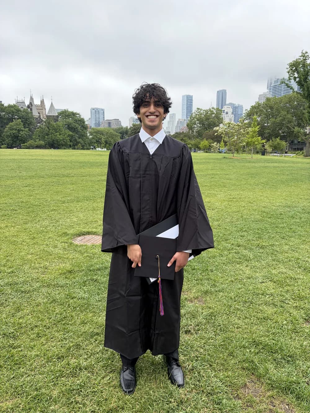 Shaan in cap and gown on a lawn with the Toronto skyline behind him
